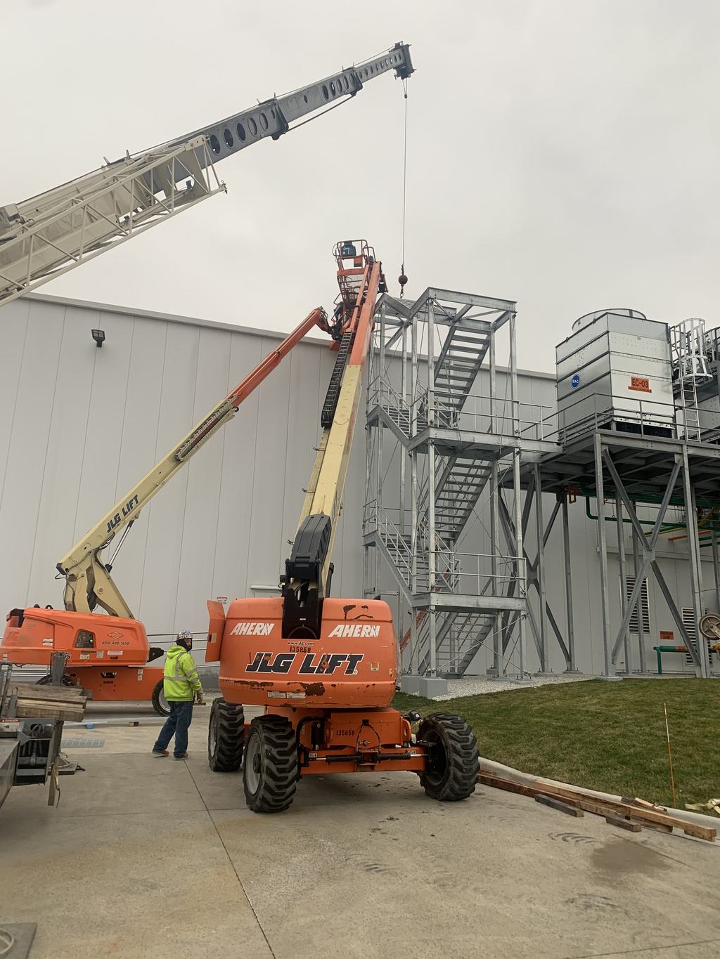 Galvanized external stair tower rising against the insulated metal panel wall beside an elevated condenser platform, with boom lifts working below