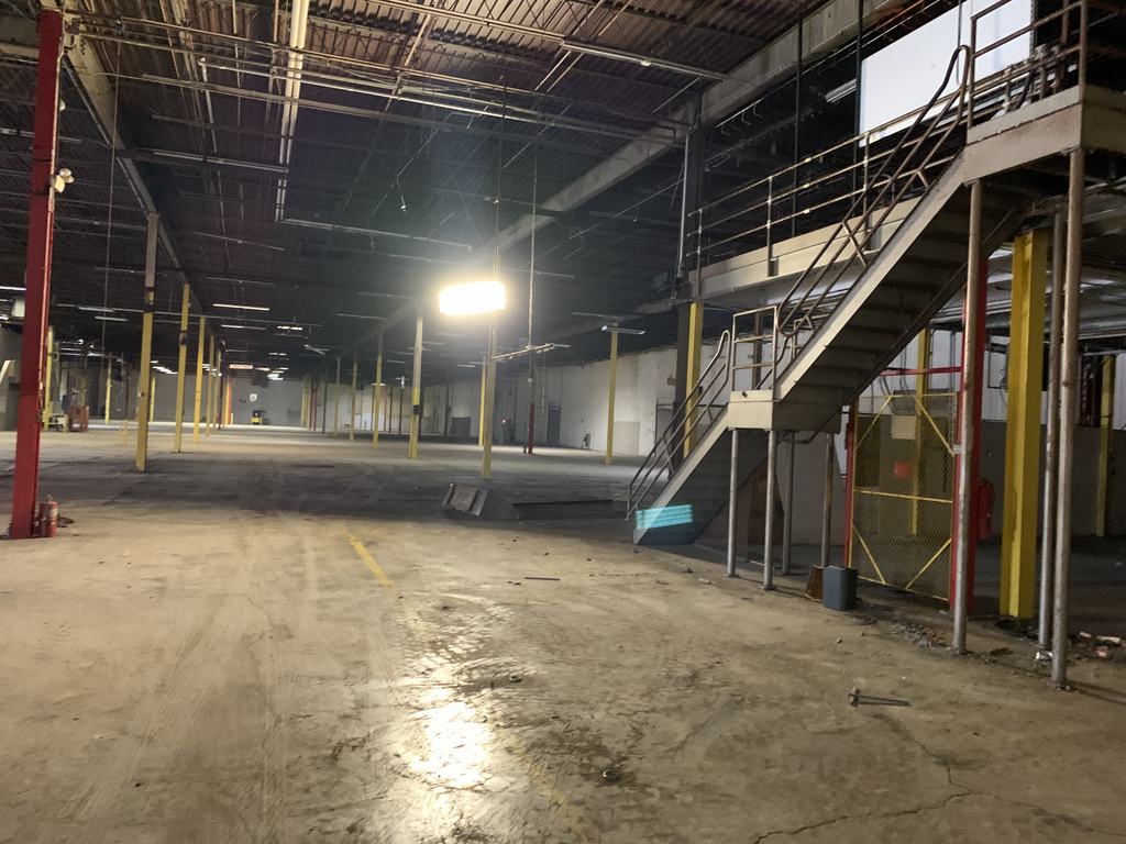Existing warehouse bay before renovation, with an open stair, exposed roof structure, and a worn floor slab