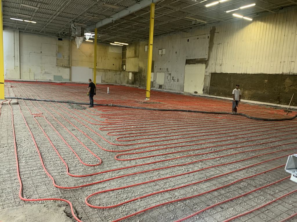 Tubing and reinforcing mesh run across the full width of a bay, with two workers laying out the pattern against the existing walls